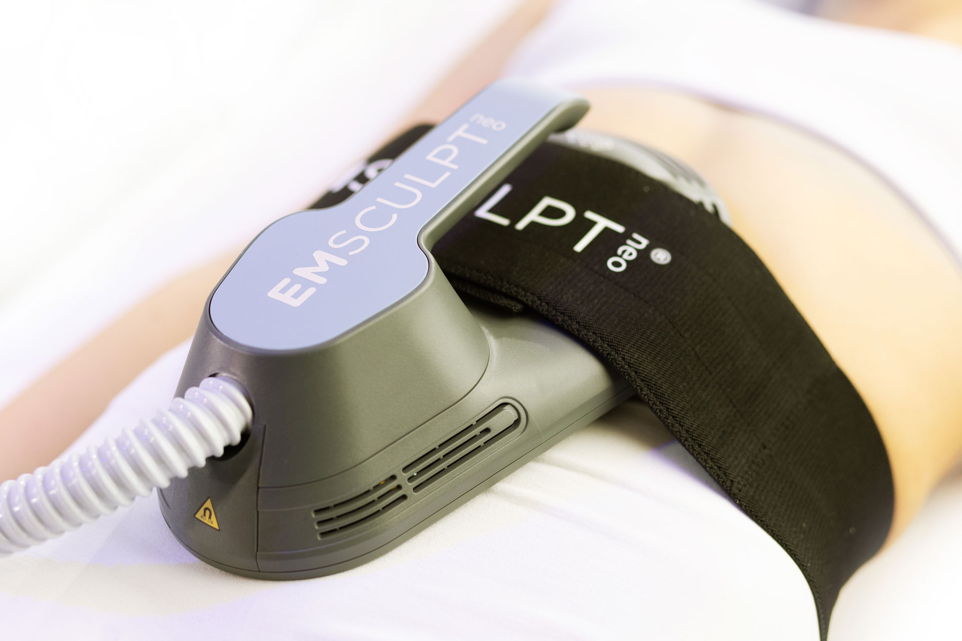 A close-up of a person's torso with an EMSCULPT NEO device strapped around the waist. The device is gray with a flexible hose connected, aiming to tone and strengthen muscles. The background is softly blurred.