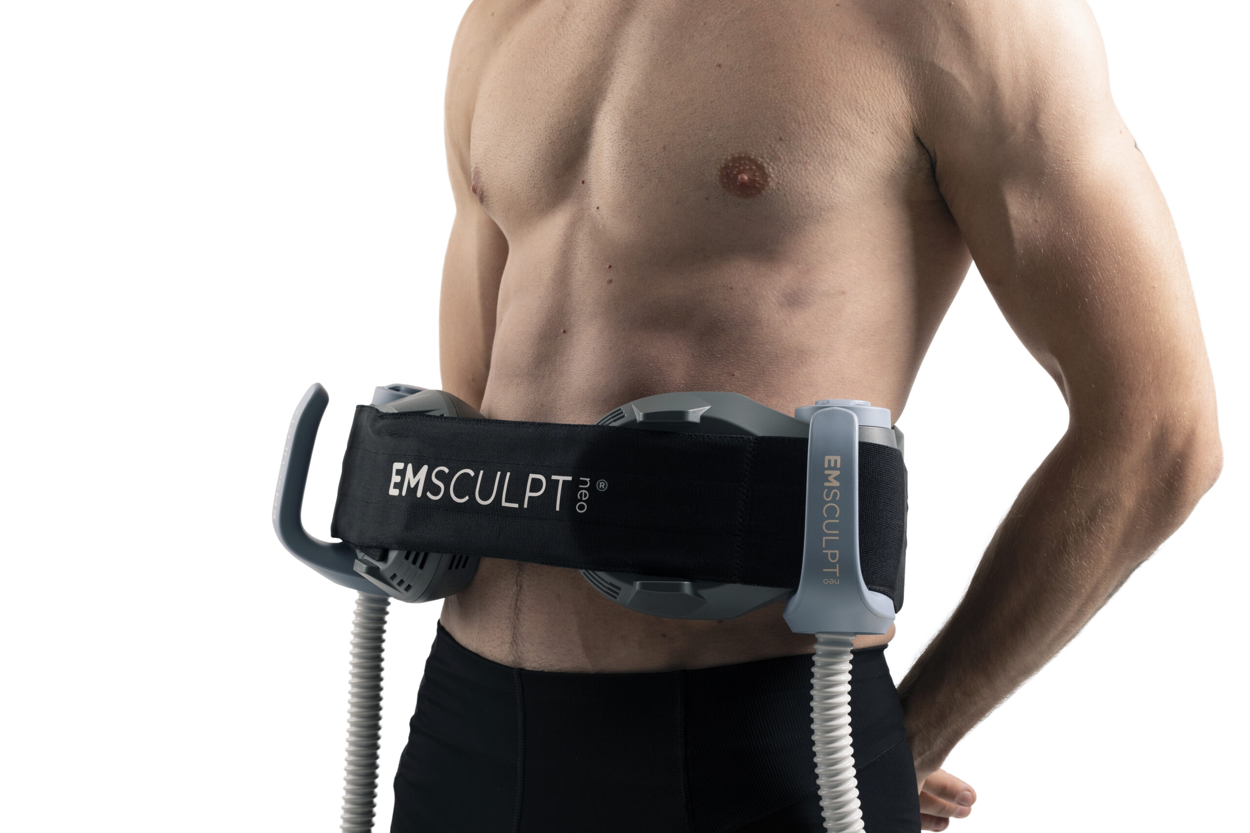 A shirtless person uses an EMSculpt Pro device on their abdomen. The device has two paddles attached with straps and cords, designed for muscle toning. The person is wearing black shorts.
