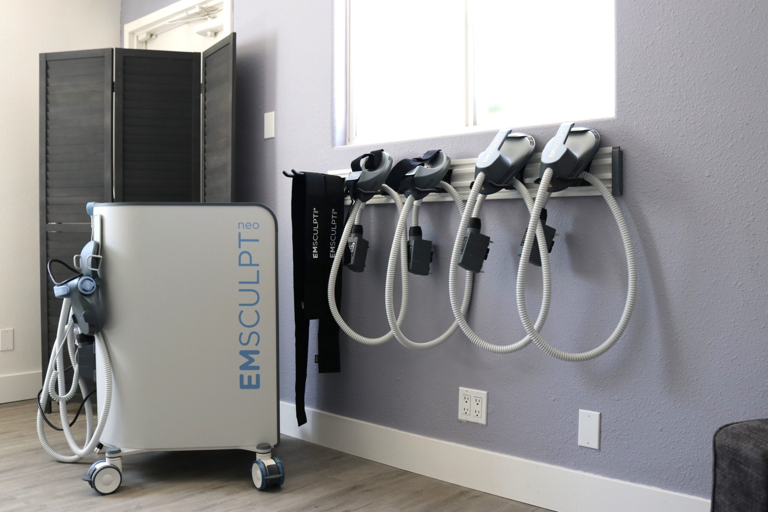 A room featuring an Emsculpt Neo machine with its attachments hanging on a wall rack. The machine is positioned on the left with a soft purple wall and a window in the background. A dark wooden partition is partially visible on the left.