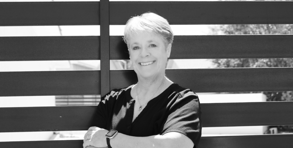 A black and white photo of a smiling person with short hair, wearing a dark short-sleeve shirt and a wristwatch. They are standing with arms crossed in front of a modern metal fence, with blurred buildings and trees in the background.