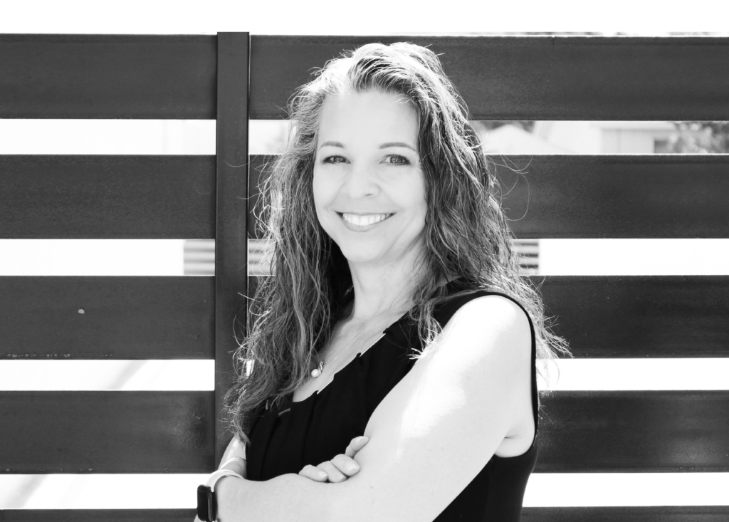 A person with long, wavy hair smiles while standing with arms crossed in front of a slatted fence. They are wearing a sleeveless top and appear confident, bathed in bright, natural light. The image is in black and white.