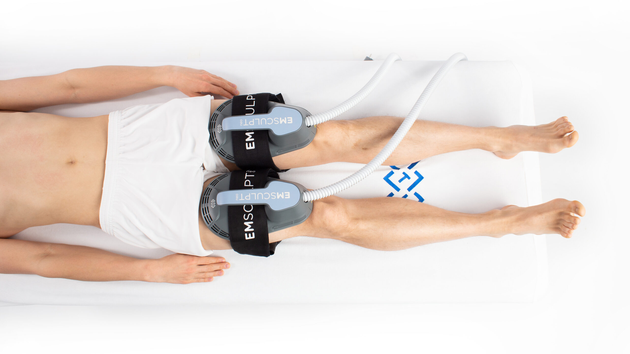 A person lying on a white surface with two devices attached to their thighs, labeled "EMSCULPT". The person is wearing white shorts, and the devices are connected by tubes extending outward.