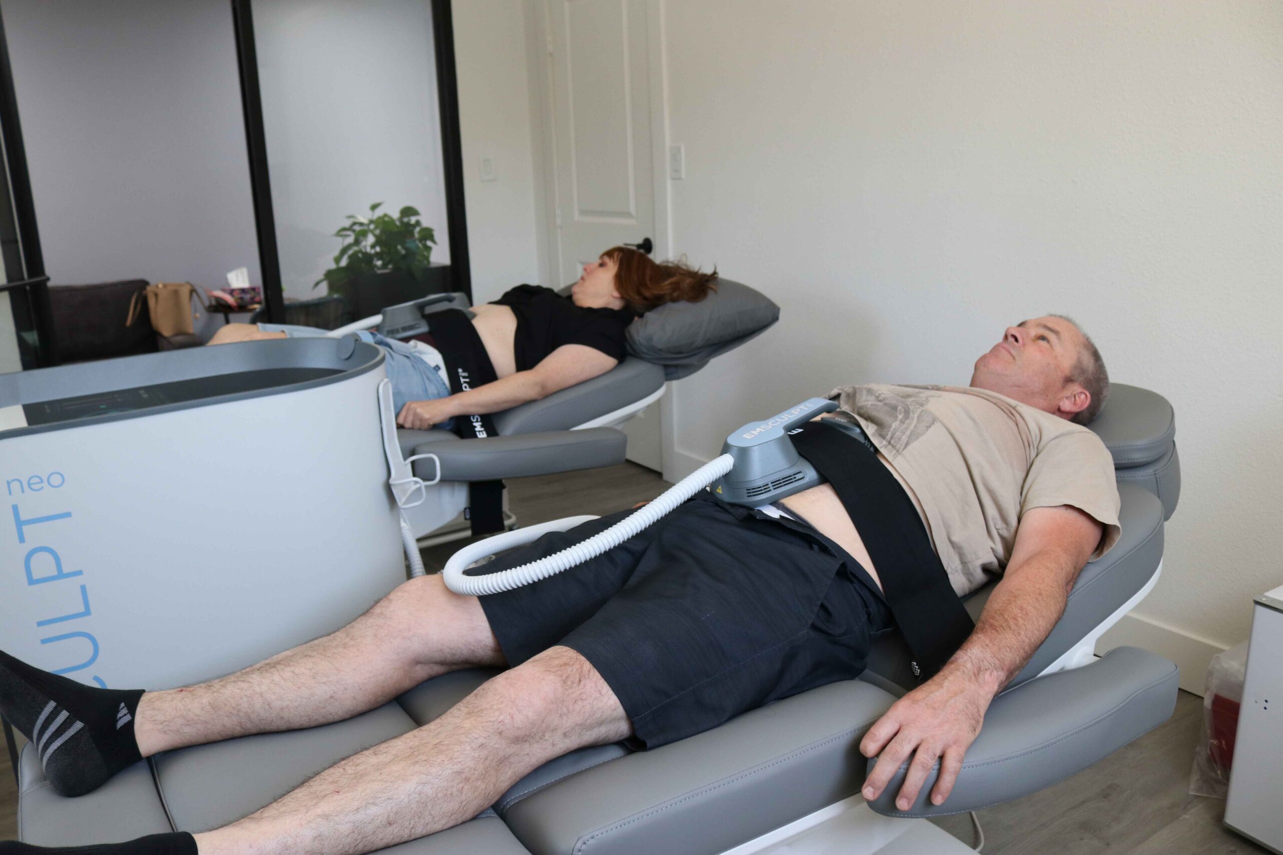 Two individuals are lying on separate chairs in a clinic, each receiving electromagnetic body sculpting treatments on their abdominal area. They appear relaxed, and the room is softly lit.