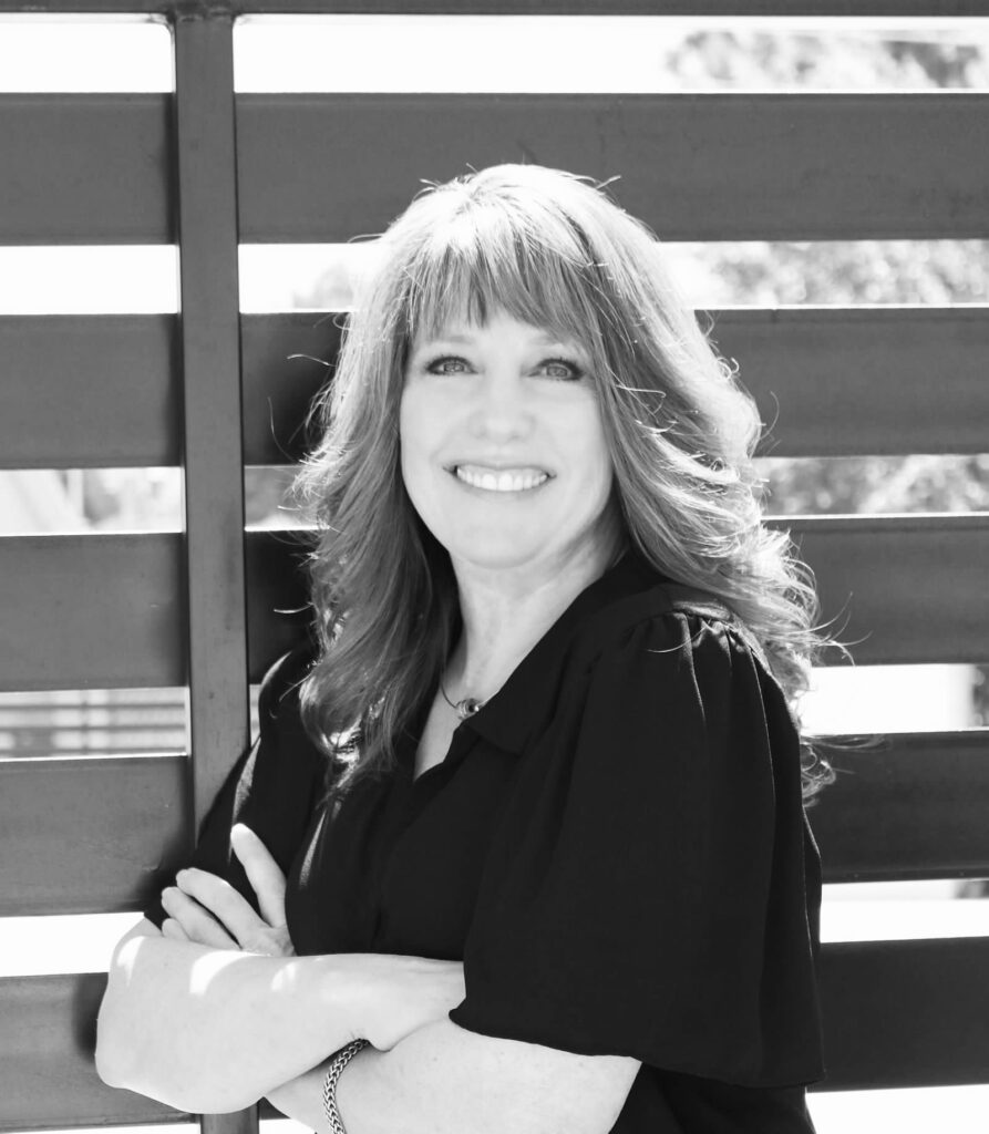 A woman with long hair smiles while standing in front of a modern slatted fence outdoors. She is wearing a black top and has her arms crossed. The image is in black and white.
